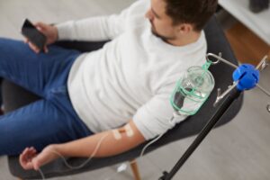 Top view of a man using a smartphone while receiving hangover IV drip therapy, replenishing hydration and vitamins for quick recovery.
